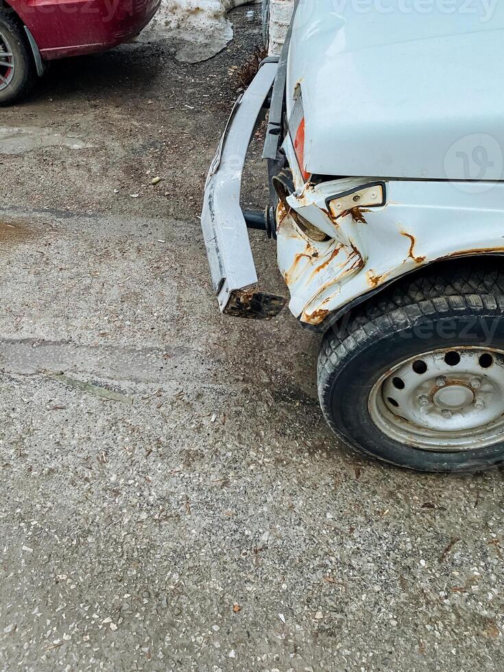 A white car that has a damaged bumper is parked on the side of the road, taking up space and possibly attracting the attention of passing motorists and pedestrians alike photo