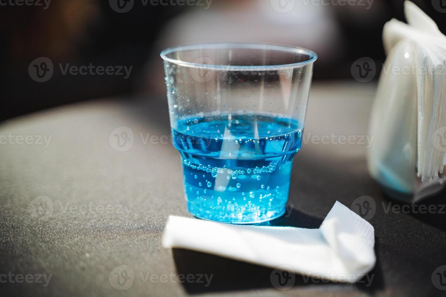 On a wooden table, a cup filled with a vibrant blue liquid is positioned next to a folded napkin, creating a colorful and inviting setting for a refreshing drink or cocktail photo