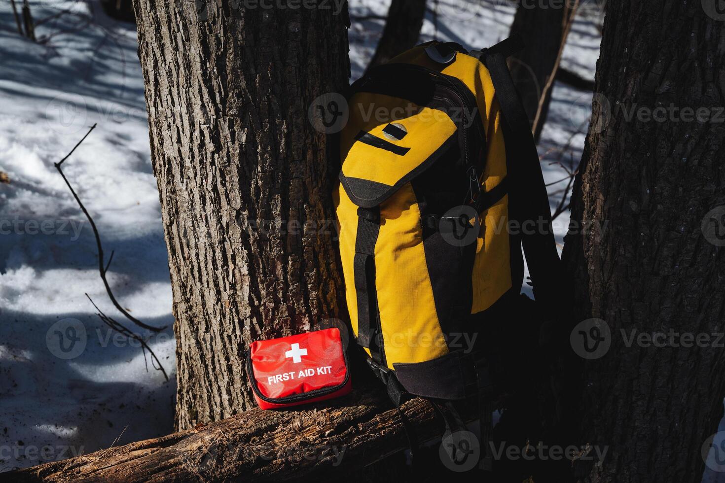 Essential Hiking Gear A bright yellow backpack with a wellstocked first aid kit is seen resting against a sturdy tree in the serene, snowy wilderness landscape photo