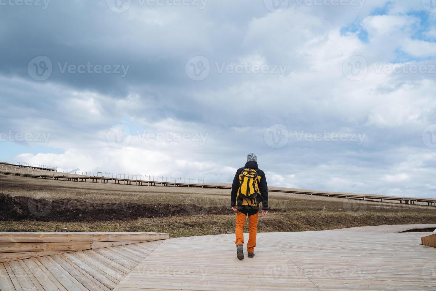 A man, equipped with a sturdy backpack, is strolling leisurely along an extended wooden walkway that gracefully winds through a beautiful and scenic area surrounded by nature photo