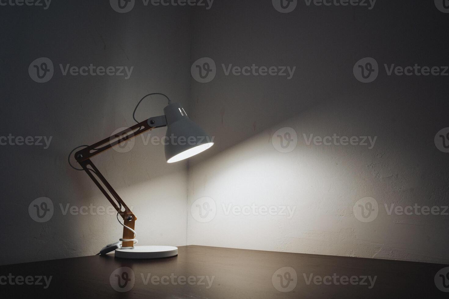 A stylish desk lamp is prominently sitting on a wooden table in an otherwise dark room, casting soft light and creating intriguing shadows that enhance the atmosphere within the space photo