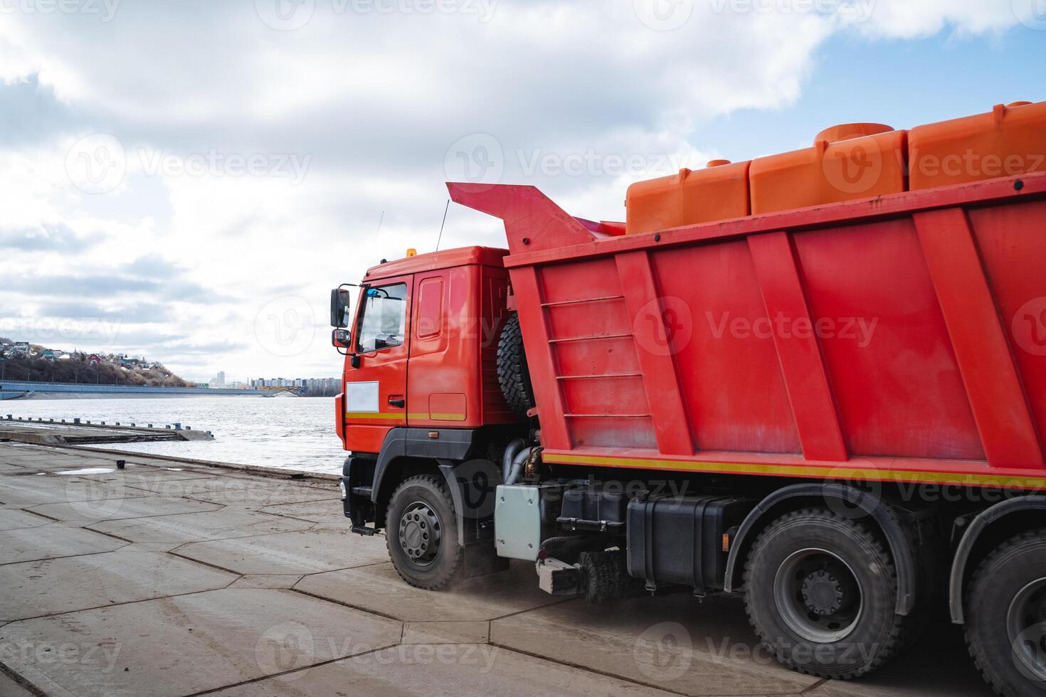 A bright red dump truck is parked alongside a serene body of water, adding a splash of color to the picturesque landscape and showcasing its robust design and functionality photo