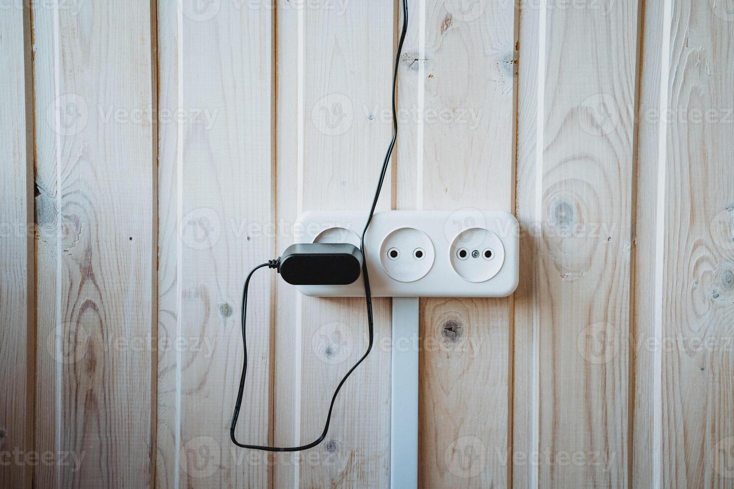 A black electrical cord is securely plugged into a white electrical outlet that is mounted on a hardwood wall made of wooden planks, providing necessary power for various devices photo