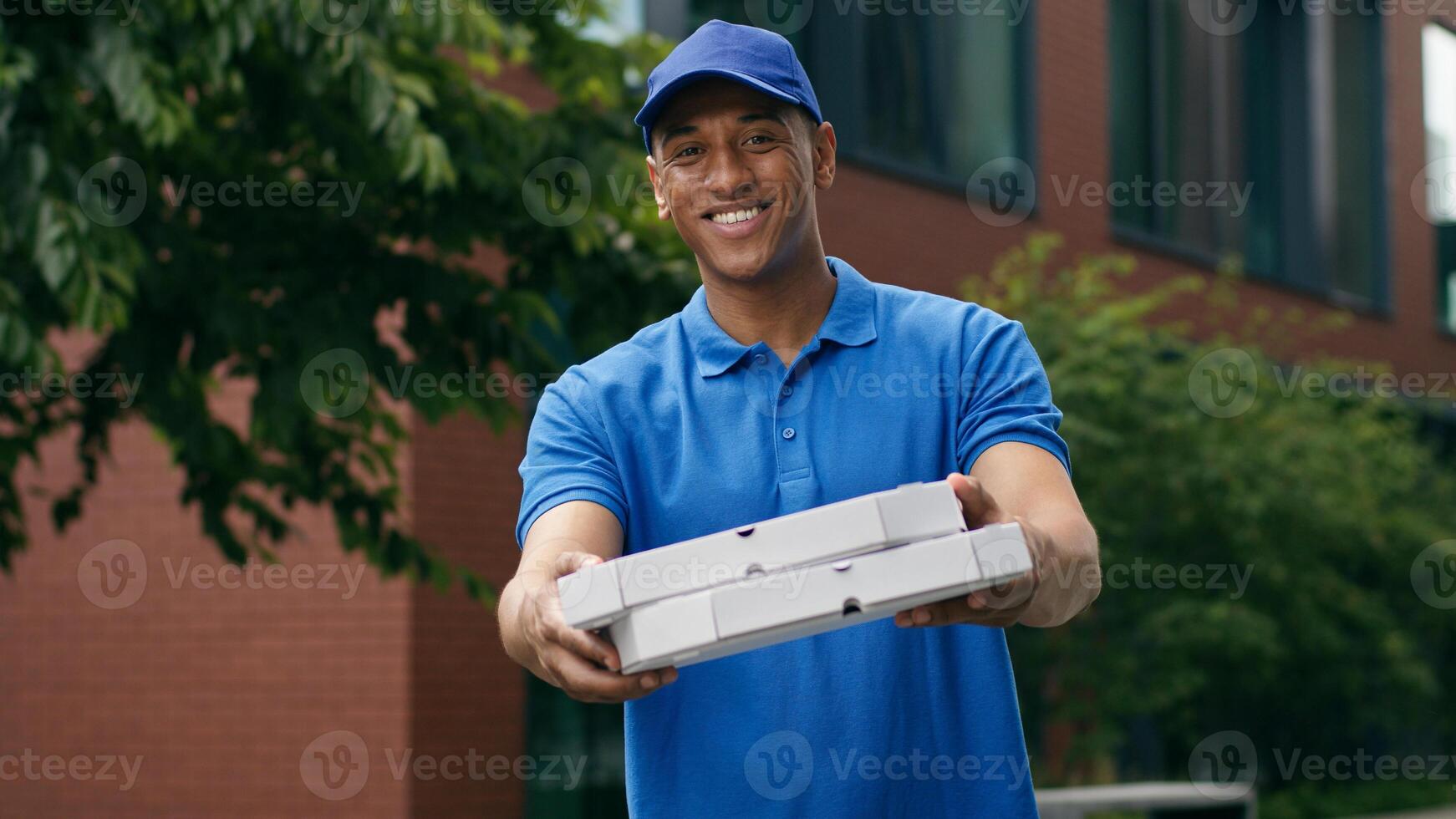Smiling male cafe pizzeria worker delivery African American man guy courier holding cardboard box parcel pizza fast food deliver meal looking at camera stand at street outdoor in city client service photo