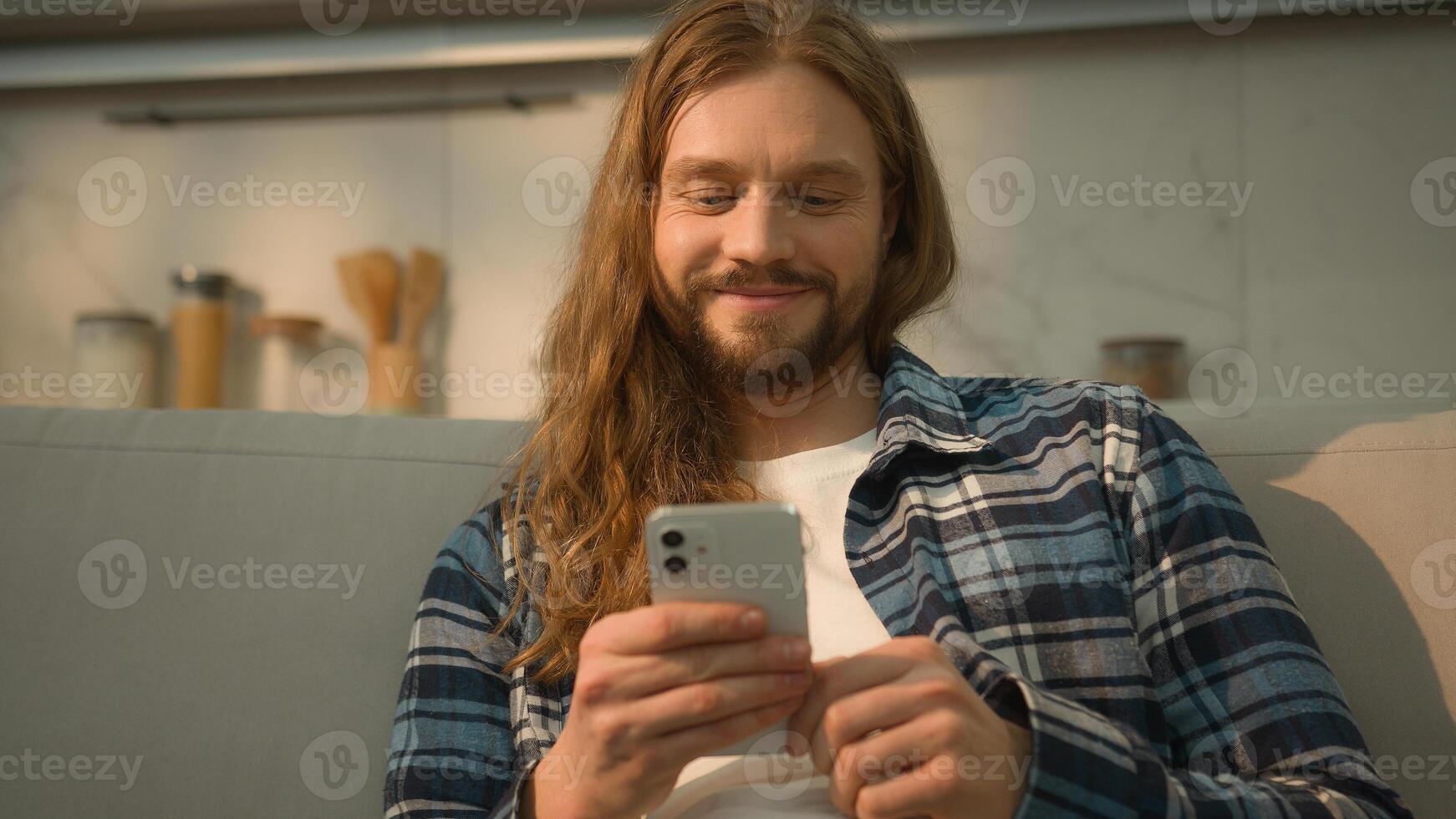 Happy smiling carefree positive relaxed Caucasian man guy male user indoors at home couch comfortable sofa using mobile phone scrolling social apps media laughing reading watching with smartphone web photo