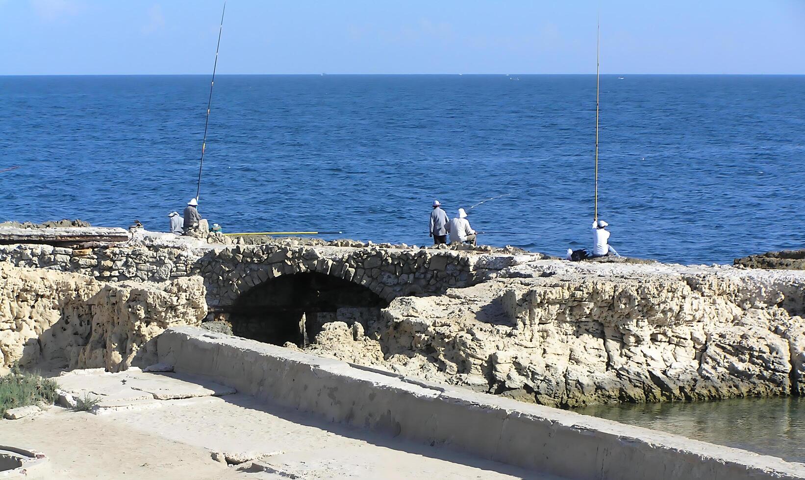 Fishermen sit on the seashore, casting their fishing rods into the calm waters. The sunlight reflects off the waves photo