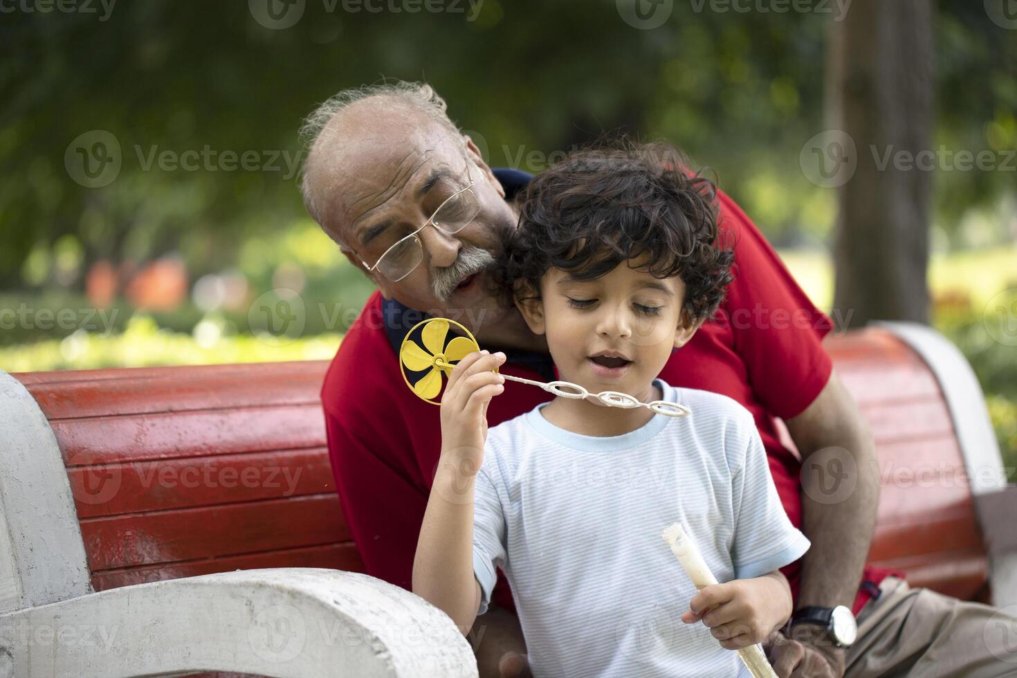 Happy Kid Blowing Soap Bubbles In Park With Grandfather While Sitting On Bench In Park photo