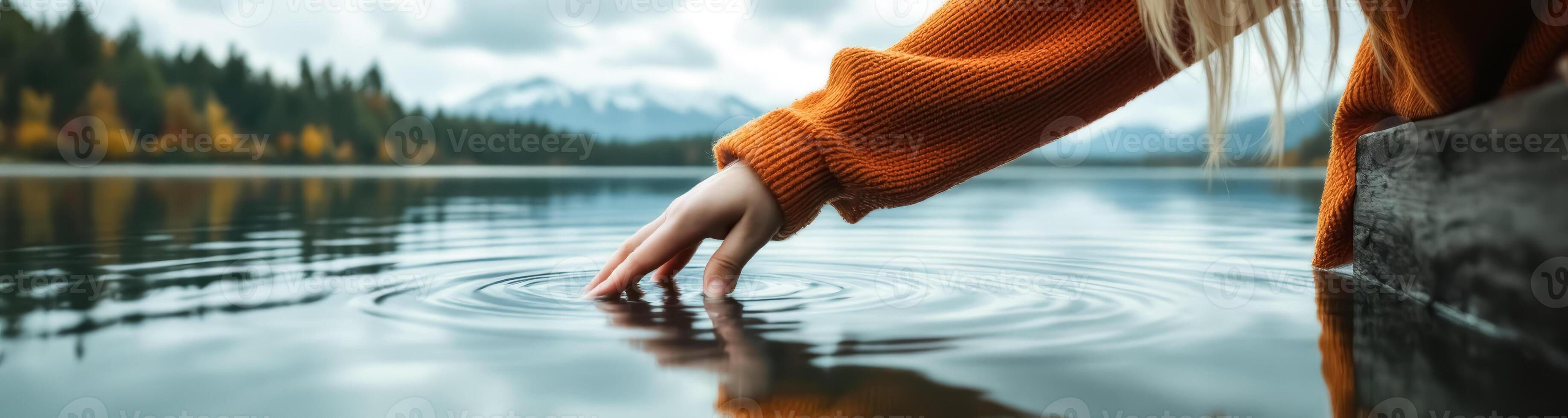 hand in a burnt sweater gently touches the surface of a calm lake photo