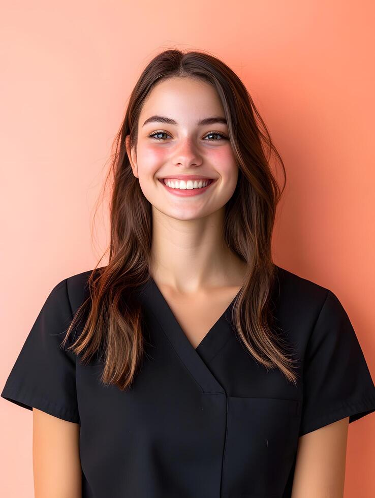 Portrait of a young friendly and confident looking hair stylist dressed in a stylish black uniform against a vibrant peach colored background that highlights her warm photo