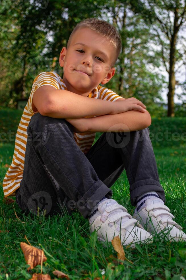 View of a schoolboy in a striped T-shirt sitting on the grass in the park photo