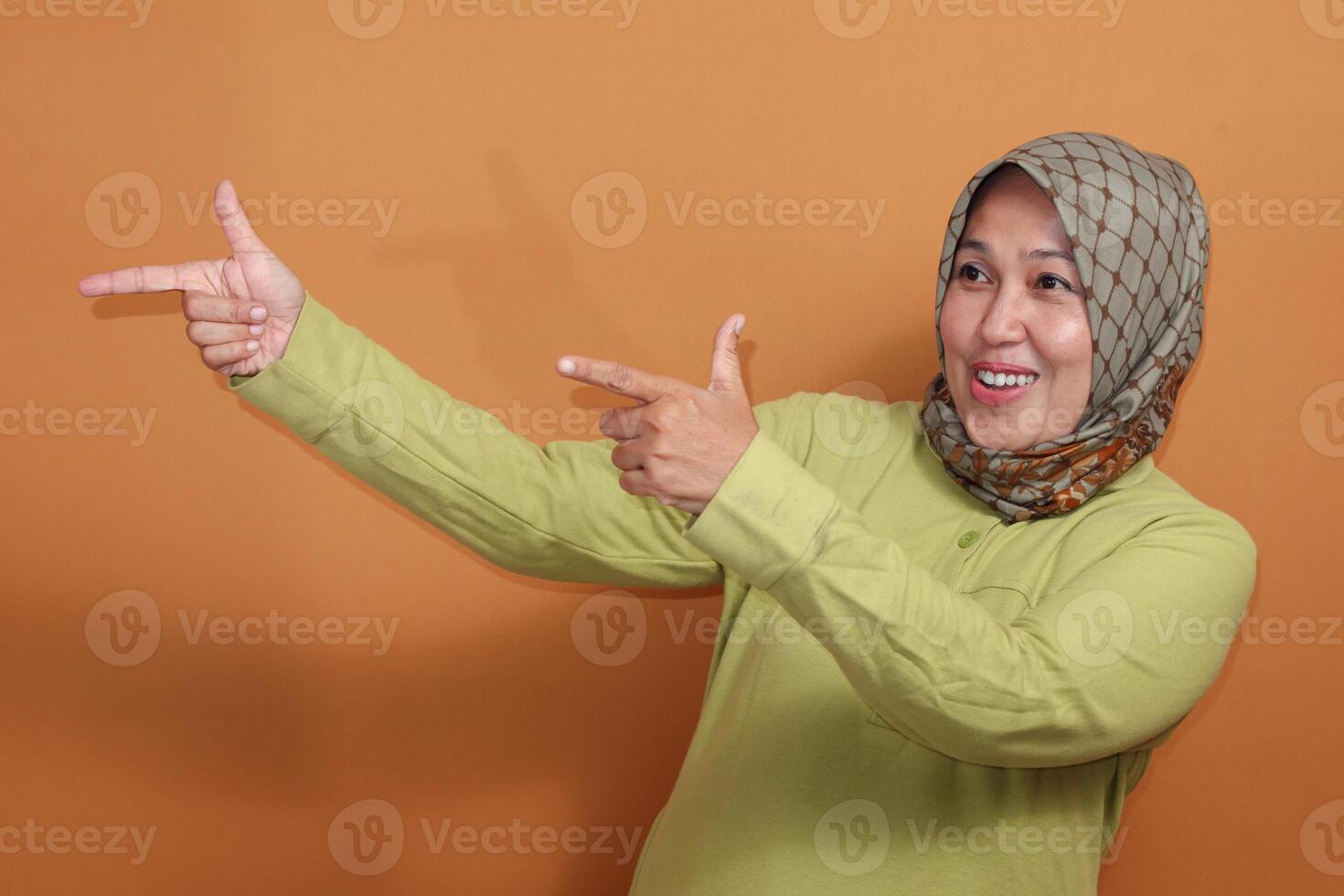 Smiling Muslim woman pointing with both hands playfully in studio, positive gesture stock photo for communication or promotion concept
