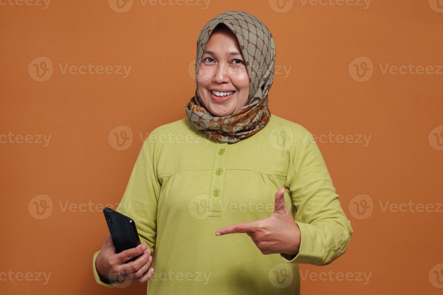 Excited Muslim woman pointing at smartphone with happy expression, ideal for digital lifestyle, technology usage, and mobile communication themes photo
