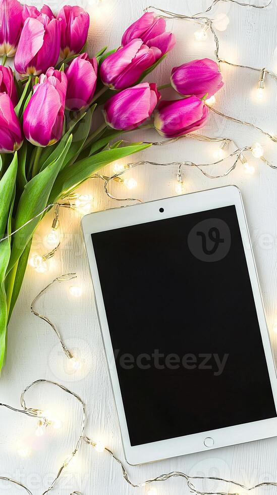 Pink Tulips and Digital Tablet on a White Surface with String Lights. Concept of Spring Decor, Modern Workspace, Technology in Nature. Mockup. Copy space photo