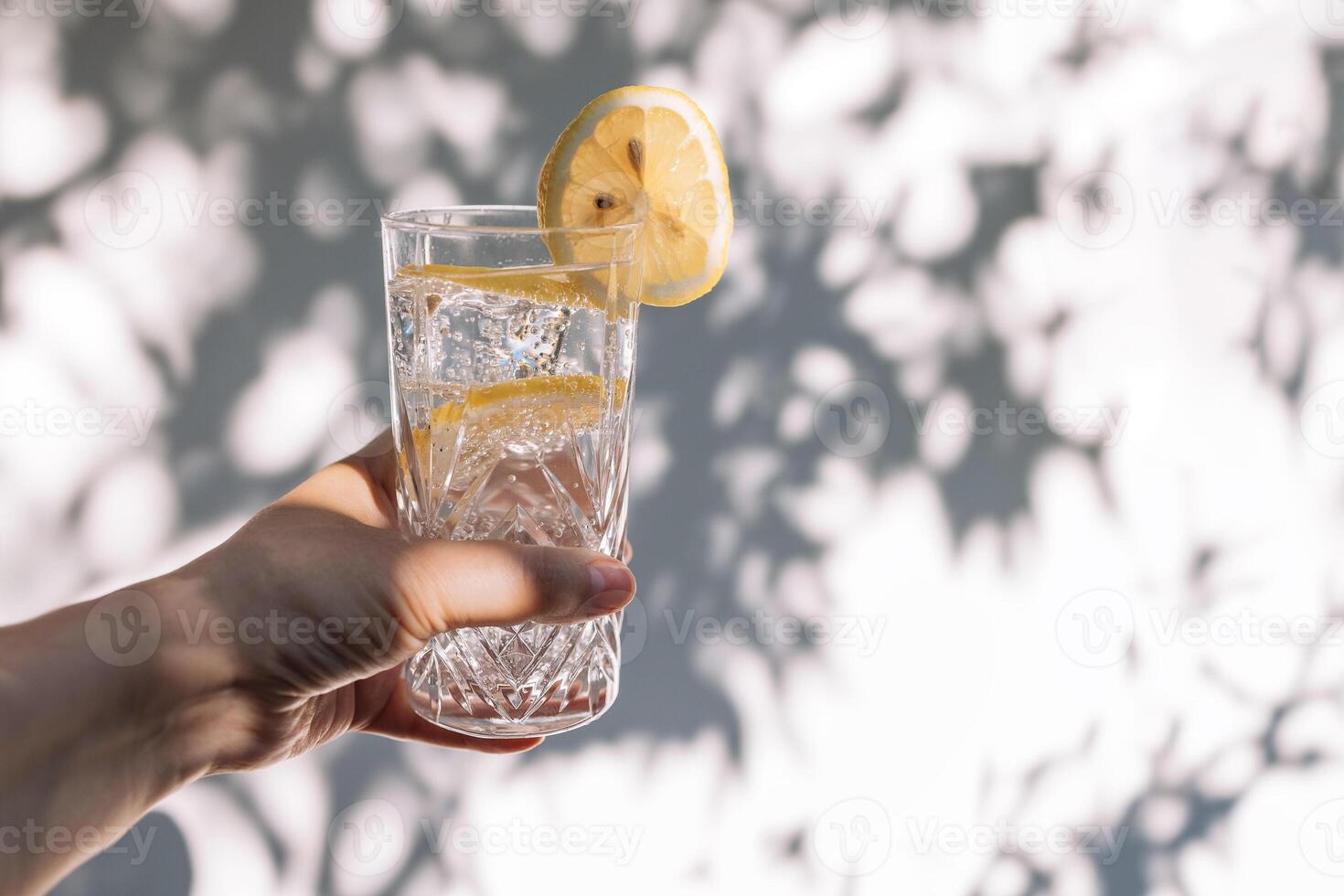 A female hand with a crystal glass with delicious cool lemonade. A woman holds an elegant cup with a sparkling citrus drink against a white wall. photo