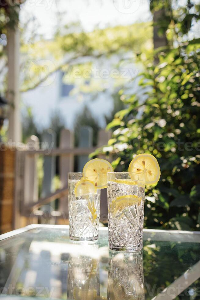 Crystal glasses with delicious cool lemonade on a glass table in the backyard of the house. Elegant cups with citrus carbonated drink. photo