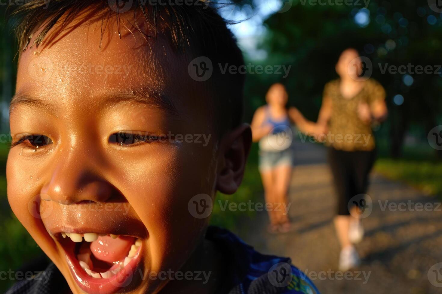 Joyful asian boy laughs exuberantly while women jog in the blurred background during golden hour. photo