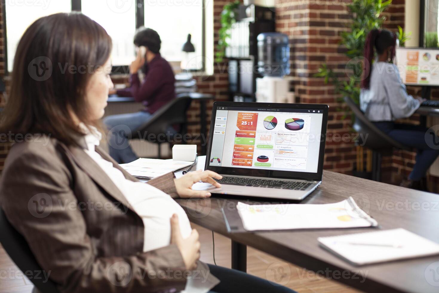 Caucasian pregnant woman with hand on stomach is reviewing company market analysis on laptop. Expectant lady using digital device at office desk, doing financial strategy research in startup office. photo