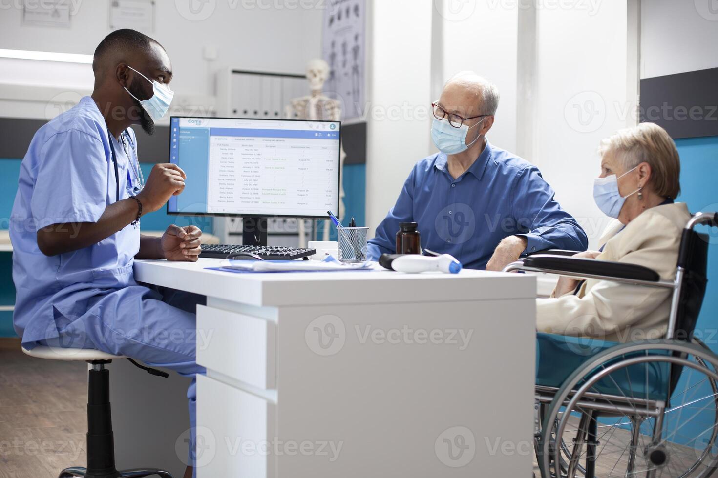 Attentive male healthcare worker uses computer to guide elderly woman in wheelchair with customized health plan. African american nurse and caucasian old couple wearing face mask in medical office. photo