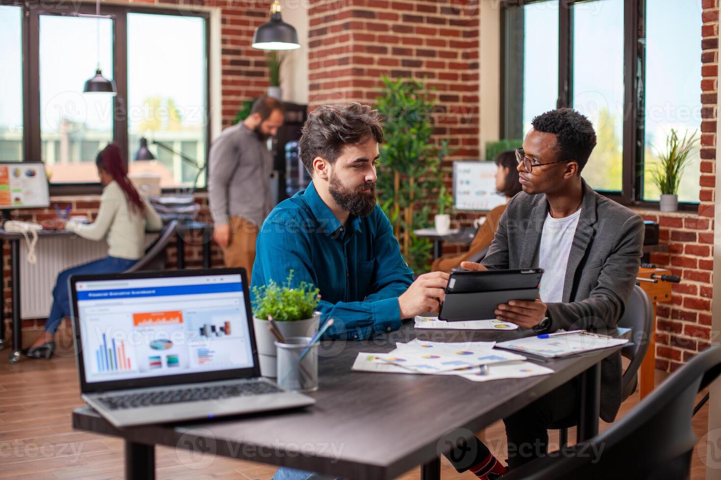 Male entrepreneurs brainstorming business ideas and reviewing company data on a tablet in modern workplace. Young men seated at desk with shared device, analyzing annual reports collaboratively. photo