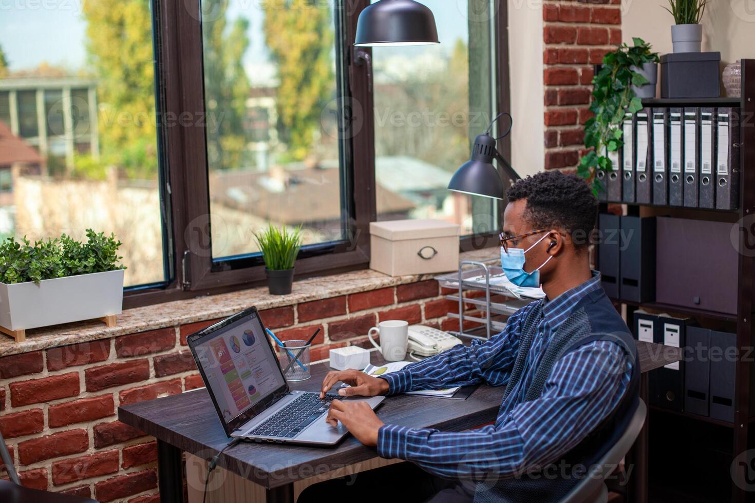 Black startup employee with face mask, wearing wireless earbuds and reviewing project insights on personal computer. Male analyst listening to music and working on laptop in brick wall office. photo