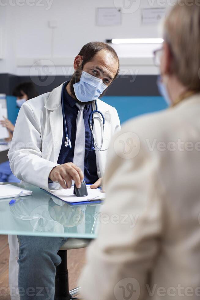 Young general practitioner in face mask is using stamper to complete prescription paper for female pensioner patient. Senior woman patiently waits as young doctor stamps medical paperwork in clinic. photo