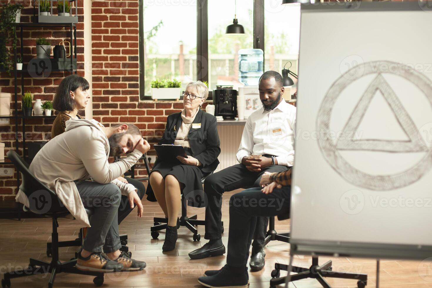 Group of alcoholics gather in aa meeting room with symbol displayed on white board. Retired therapist conducting supportive conversation, discussing mental health and addiction in brick wall office. photo