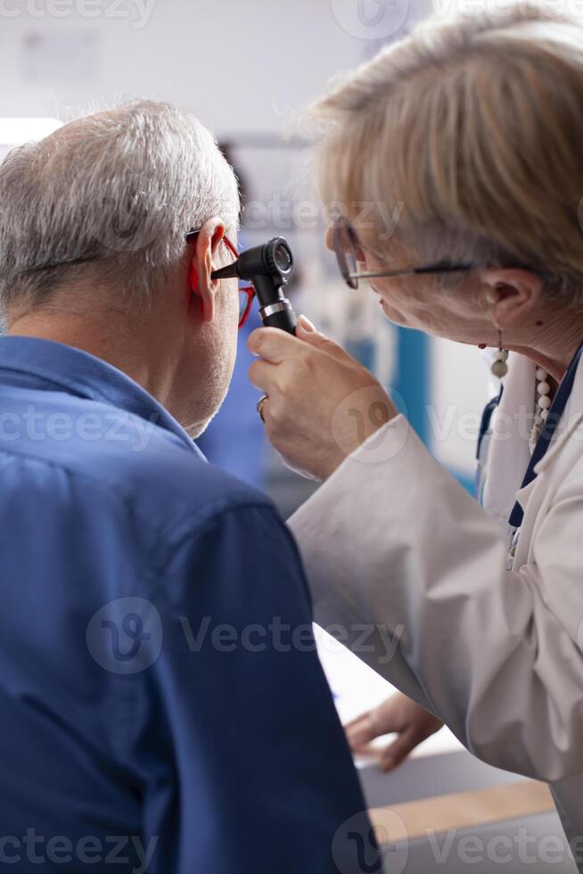 Experienced retired female physician inspecting ear of elderly male patient in clinic office. Closeup of skilled old doctor using otoscope during medical consultation of senior white man. photo