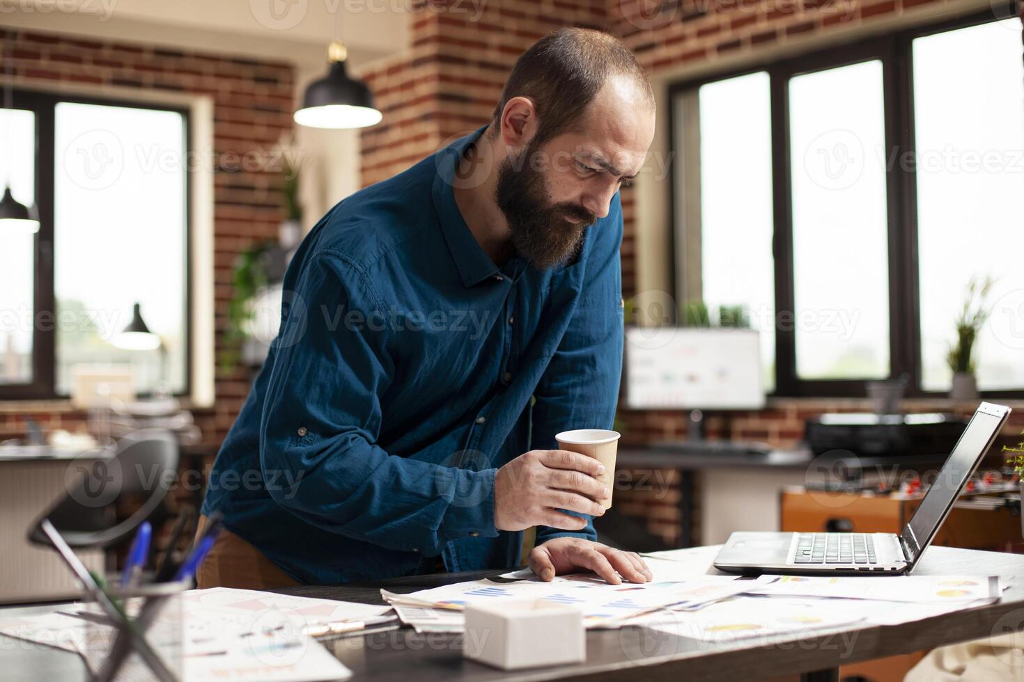 Male analyst holding coffee cup and reviewing financial paperwork on office desk in modern workspace. Professional bearded man focusing on company research documents placed on table with laptop. photo