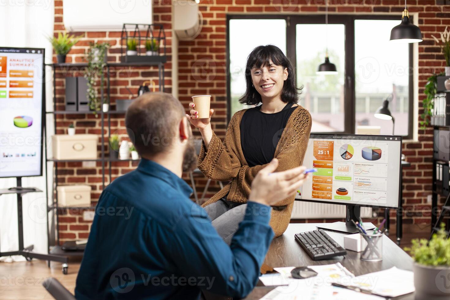 Smiling businesswoman with coffee, sitting on office desk and discussing with male coworker in brick wall room. Caucasian colleagues taking a break and having conversation in startup office. photo