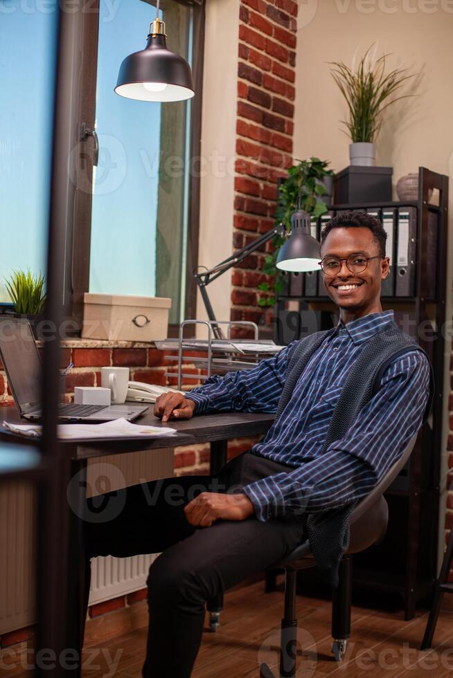 Portrait of african american man sitting at office desk and looking at camera. Black male entrepreneur with spectacles smiling brightly in home workspace with table arranged with laptop and documents. photo