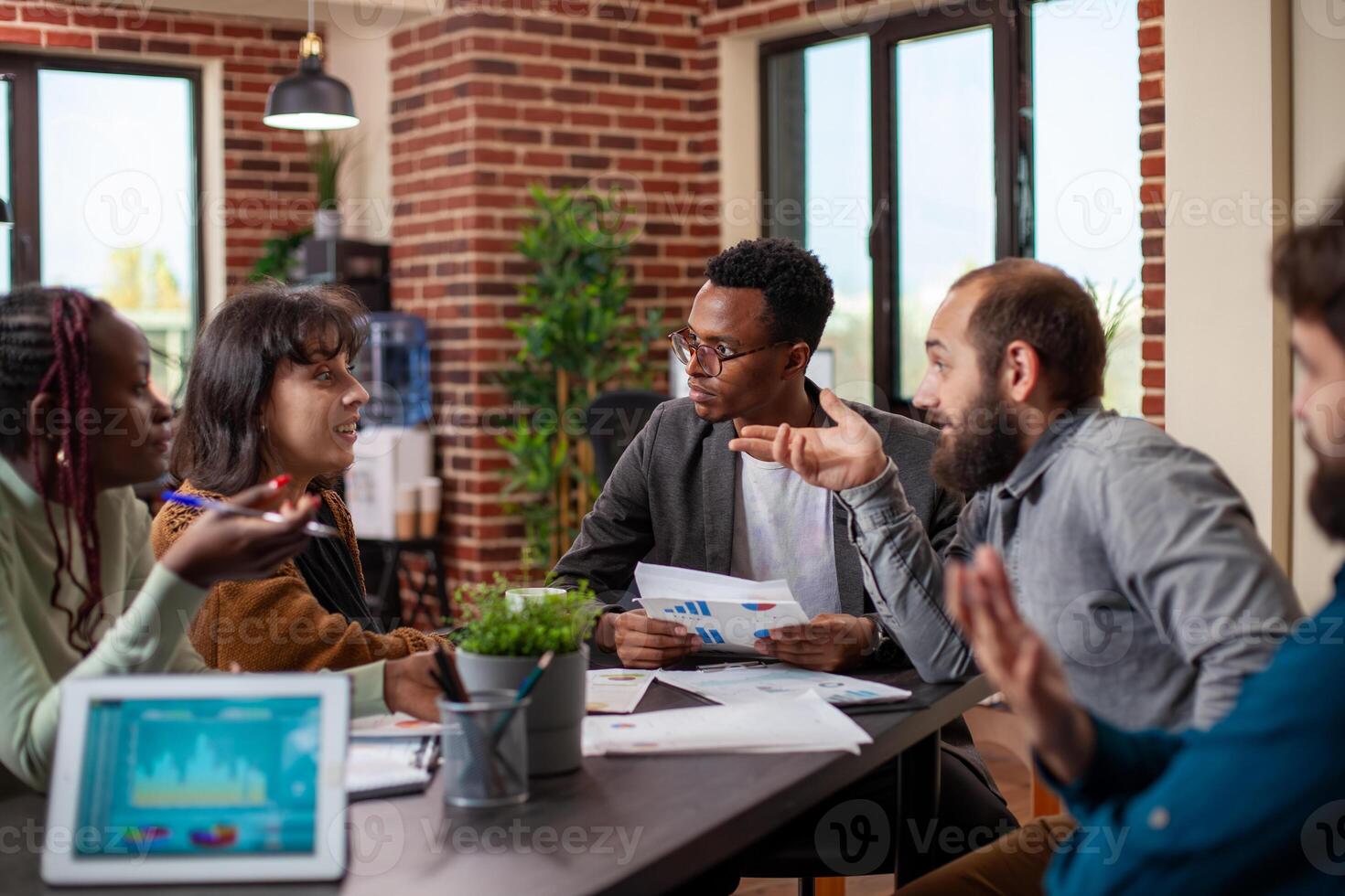 African american male manager conducting group discussion with his employees in startup office. Colleagues sitting together in brick wall workspace, having a business meeting with documents on table. photo