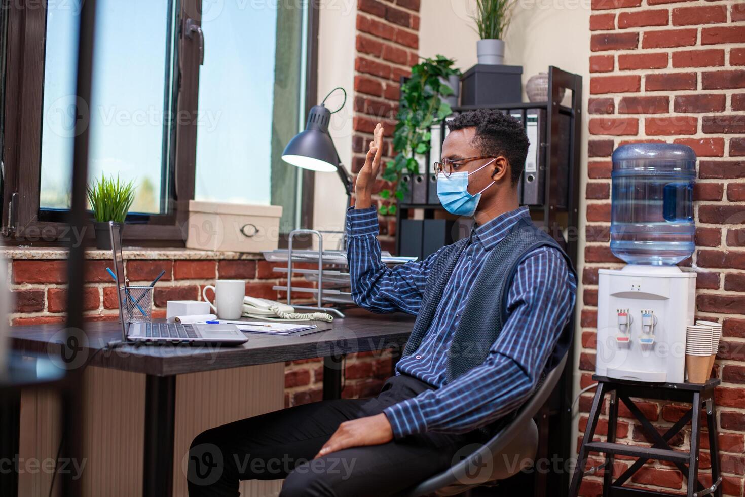 Professional advisor with face mask, sitting at table with laptop, begins videoconference with clients. Black manager waves hello and uses technology to share information online with remote workers. photo
