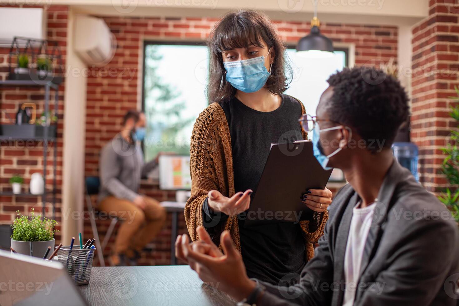 Caucasian female manager holding clipboard and advising african american male employee in startup office. Colleagues wearing face masks share ideas on business strategy in modern workspace. photo