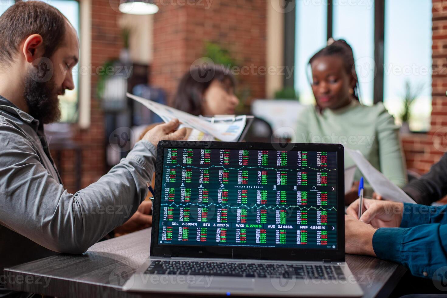Colleagues discuss in brick wall office, reviewing documents as the laptop on table displays financial statistics. Digital device on desk showing market analysis data in modern workspace. photo