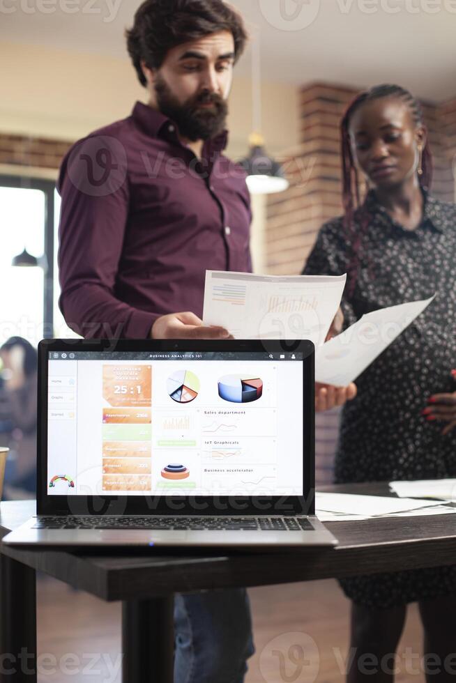 Pregnant lady and male colleague prepare market strategy as digital device shows data charts for research. Selective focus on laptop displaying financial analysis as diverse coworkers review documents photo