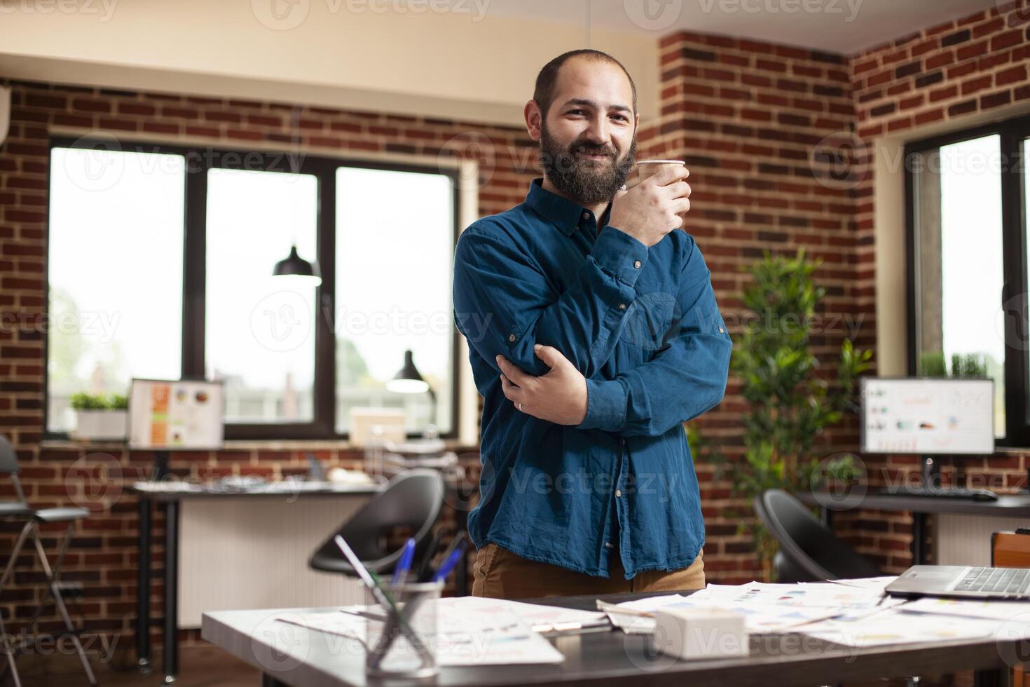 Young businessman holding coffee cup and smiling towards camera prepared to work startup office. Portrait of male analyst standing in brick wall workspace surrounded by company research documents. photo