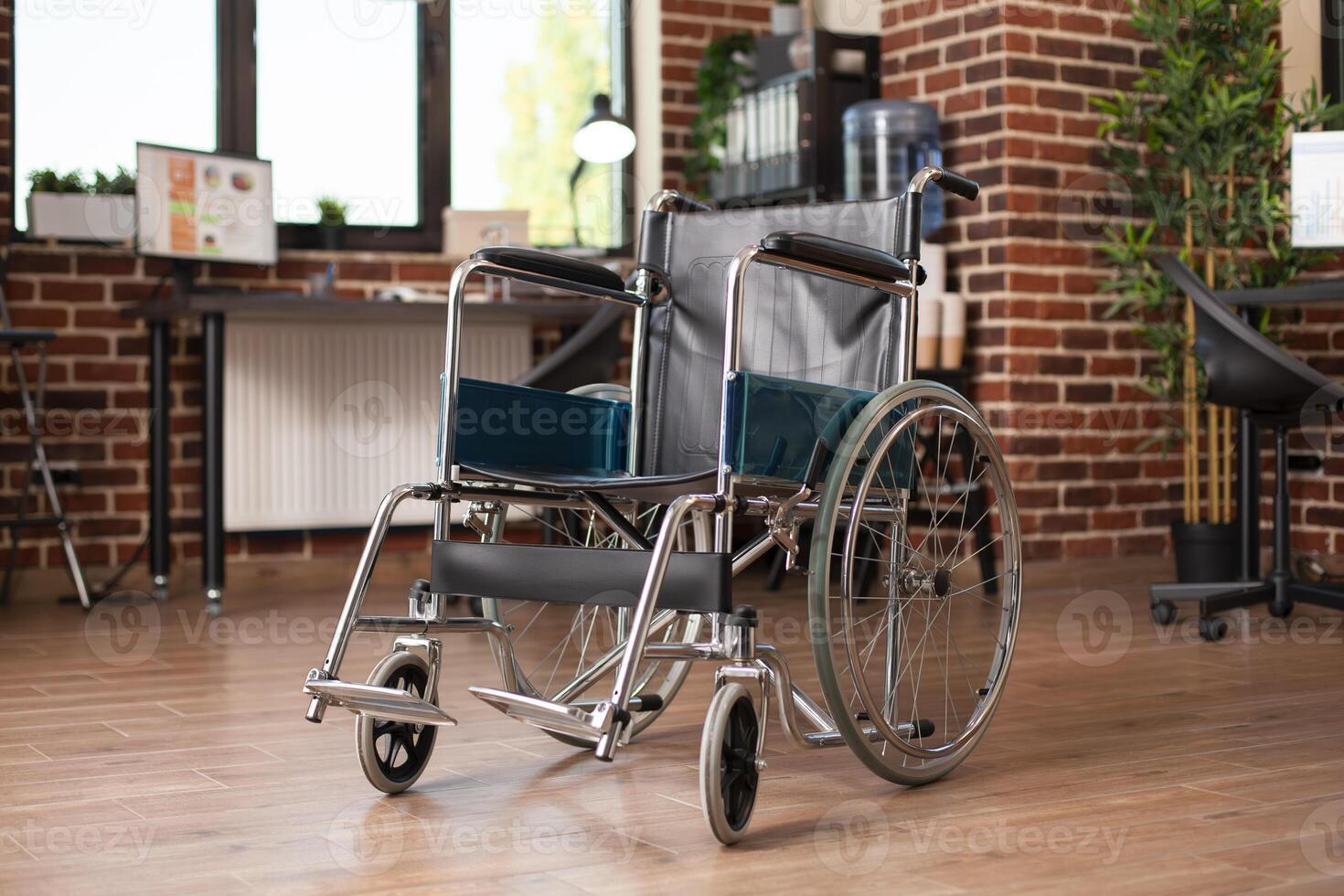 Closeup of wheelchair placed in middle of vacant workspace, with computer and business documents on desk in background. Shelves and plants complementing the spacious, organized brick wall office. photo