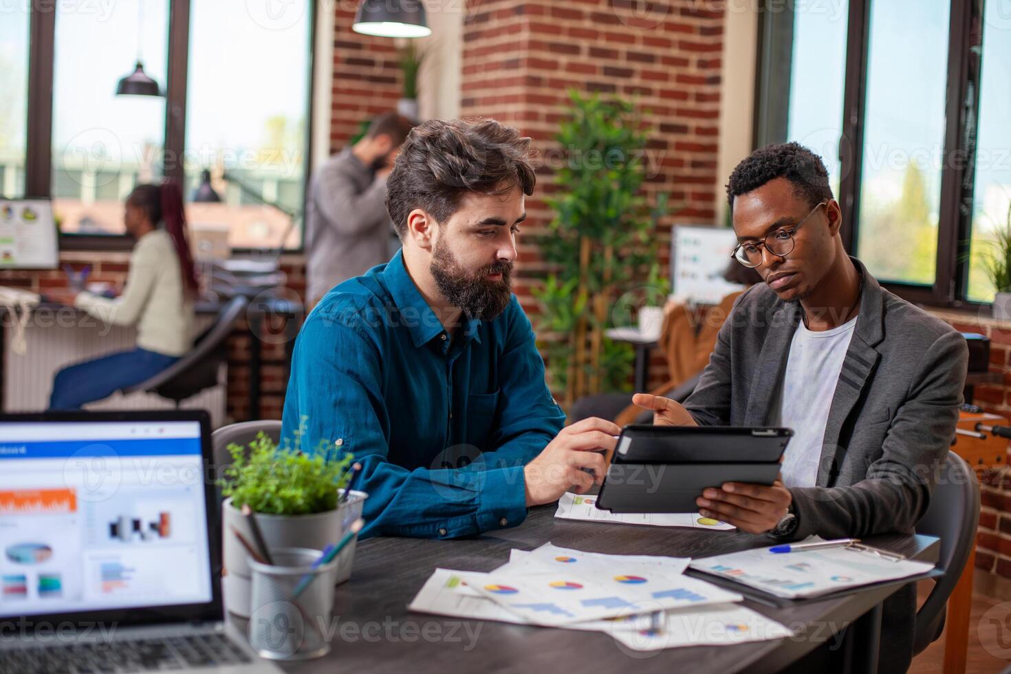 Caucasian man and black coworker analyze financial reports using tablet and documents, discussing strategy ideas. Business partners use digital device in planning and collaboration for company growth. photo