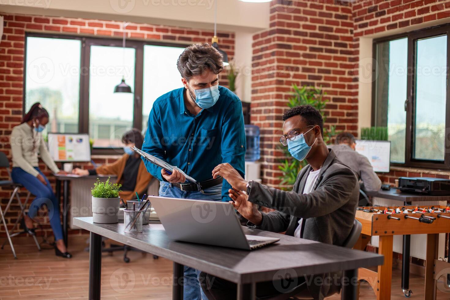 Caucasian man and African American coworker review online documents on a laptop at a table in modern workspace. Male colleagues with face masks, focus on achieving business goals at startup company. photo