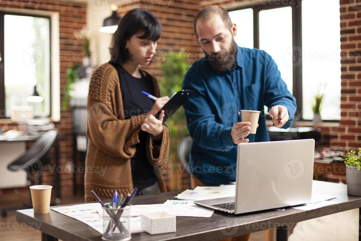 Male manager stands at desk with female coworker, using laptop to explain market analysis in startup office. Businesswoman writing on clipboard as bearded man with cup describes company research data. photo