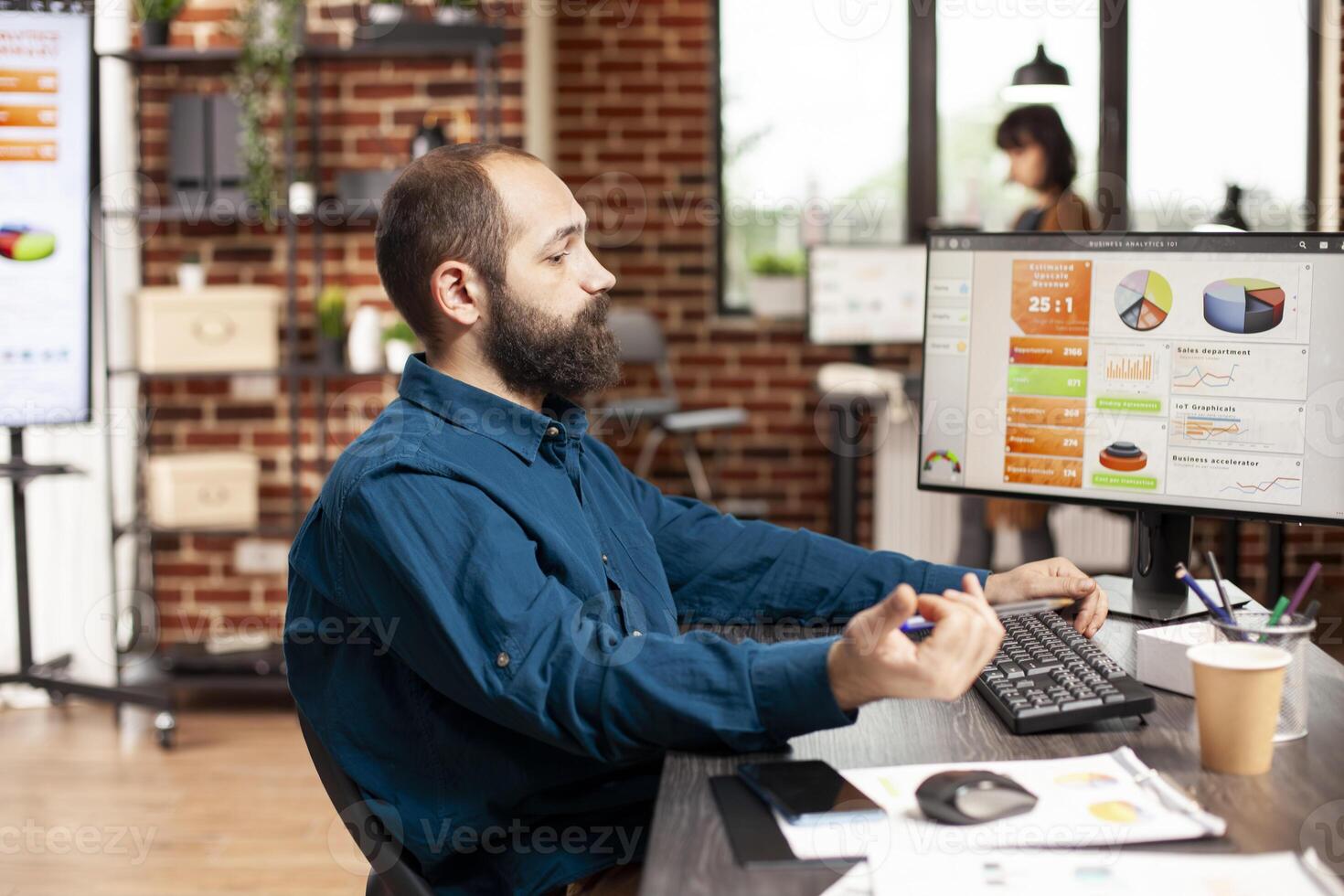 Bearded man in business casual sits at office desk, analyzing financial charts on desktop monitor. Male analyst focused on researching project ideas for startup company at brick wall workspace. photo