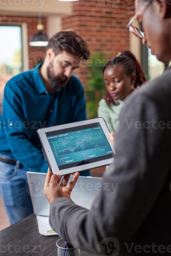 Black analyst uses tablet to assess business reports as his coworkers work together in startup office. African American manager reviews company progress on digital device, colleagues discuss nearby. photo