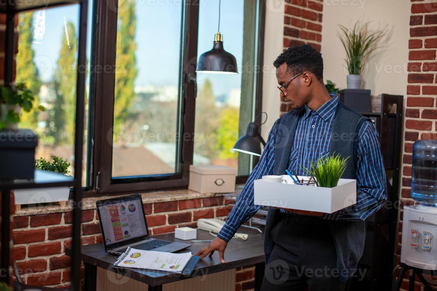 Male employee stands in workspace, holding box containing green plant and office materials, reviewing financial documents on desk. Black businessman checking company paperwork before leaving work. photo