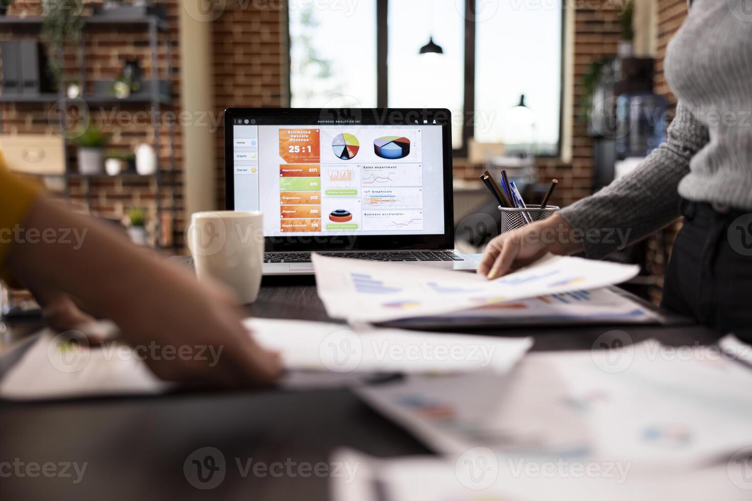 Selective focus on laptop displaying business analytics as hands of female individuals hold documents at desk. Closeup of people grasping papers over a table as digital device shows data graphs. photo