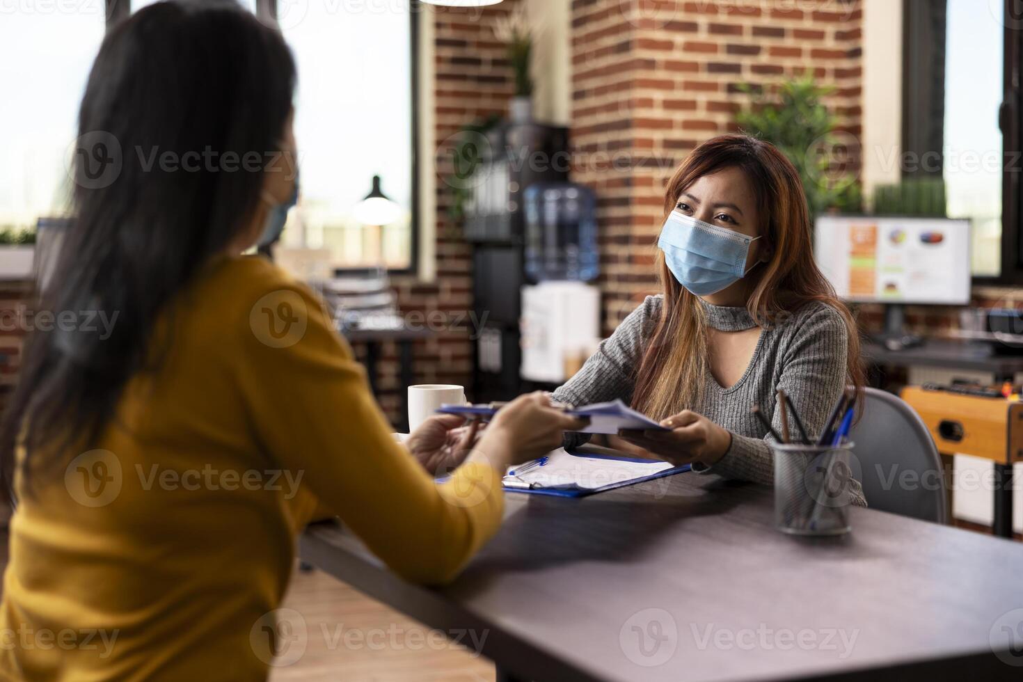 Businesswomen seated at desk, holding a clipboard and sharing strategy ideas in brick wall workspace. Asian female manager with a face mask, receiving a document from employee in startup office. photo