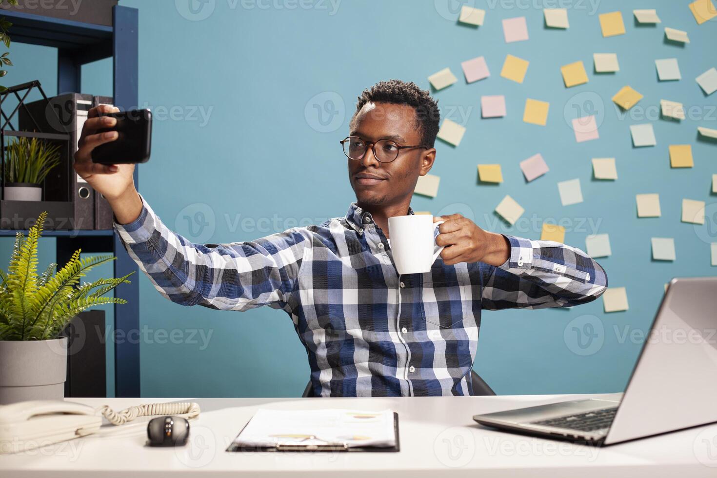 African american company advisor taking selfie photo and having a cup of tea in his modern workspace. Black businessman with cellphone is taking a of himself about to drink coffee at his desk.