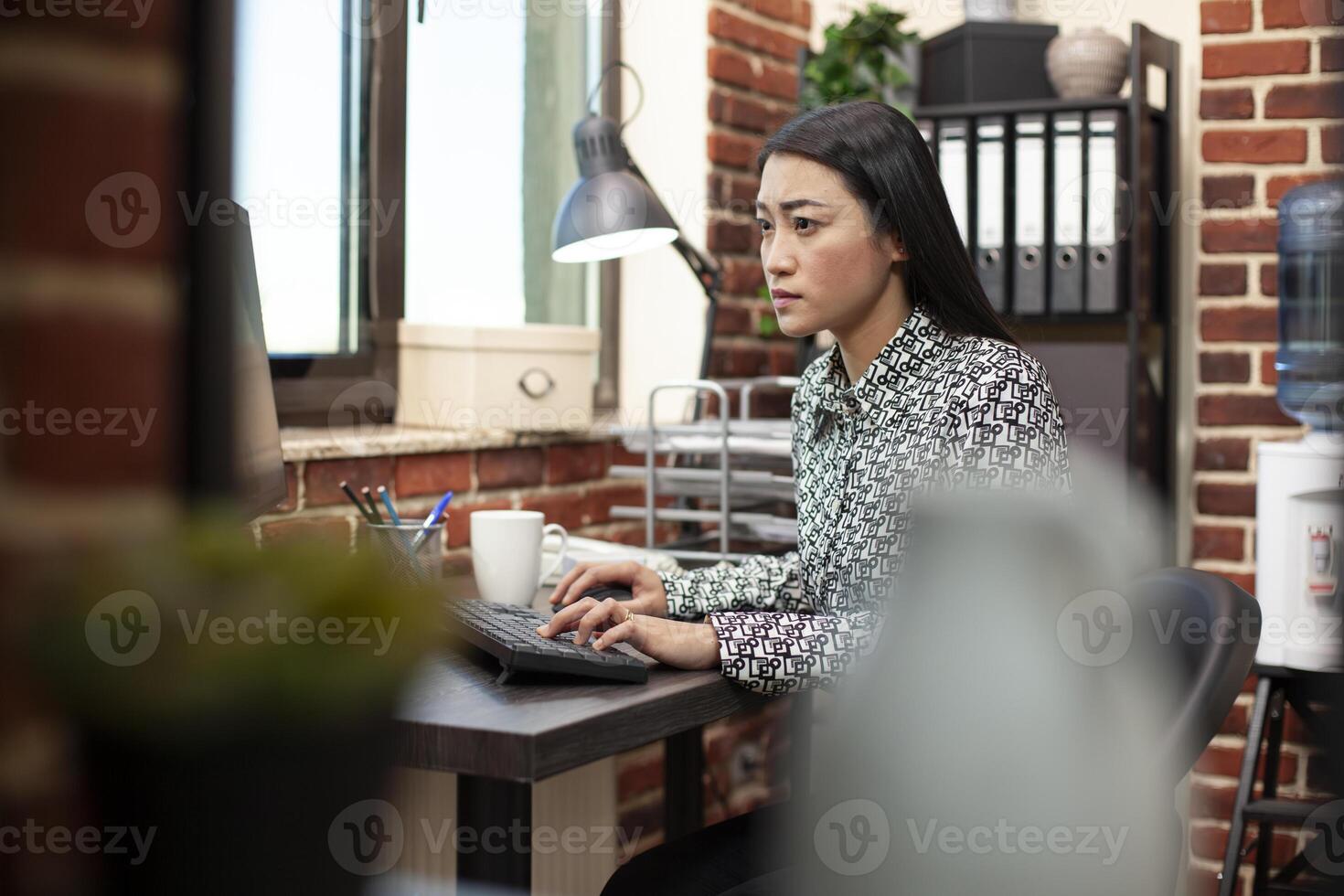 Asian woman seated at desk near window is concentrating on her work, typing and updating marketing data in startup office. Female researcher using her computer in brick wall workspace. photo
