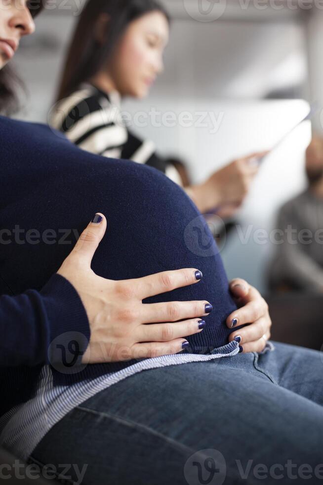 Closeup of a white woman embracing her pregnancy belly at job interview appointment, waiting in line. Expectant lady touching her baby bump and sitting in queue for meeting with hr department. photo