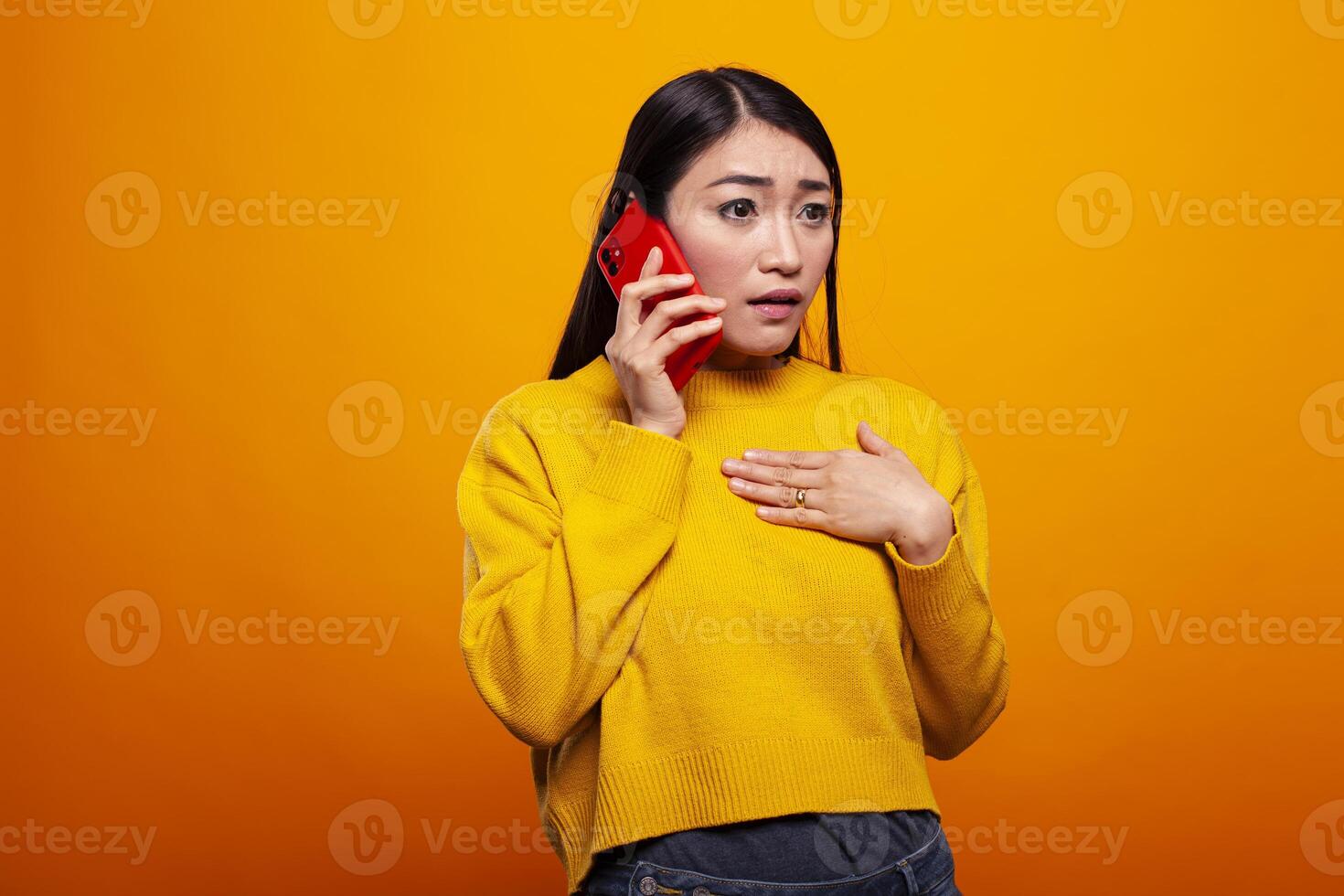 Woman holding her mobile device to her ear and having a phone conversation, receiving shocking information. Asian lady standing with hand to chest and looking startled by bad news from a call. photo