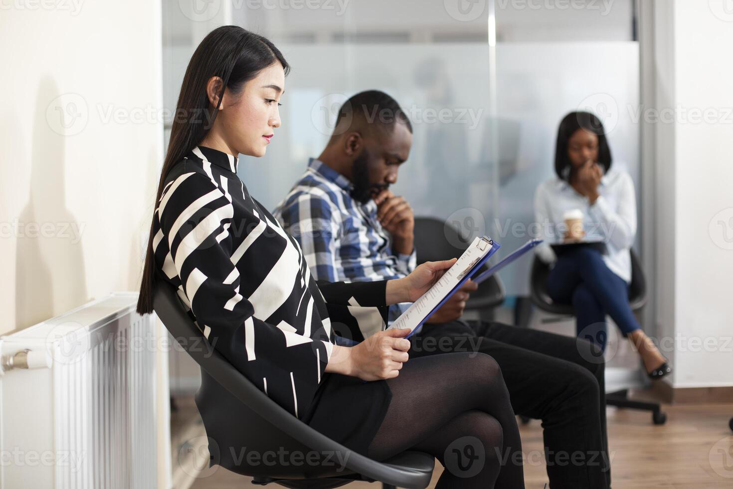 Unemployed woman sits on chair, reviewing her cv resume and waiting with other applicants. Asian female candidate checks job details on clipboard, sitting in line with black people in office lobby. photo